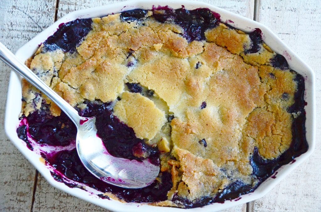 Blueberry cobbler with golden top and a spoon on a white wood table, Maureen ABood