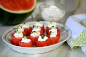 Watermelon bites with labneh and cucumber on a plate, sourrounded by watermelon and a green-trimmed towel, Maureen abood