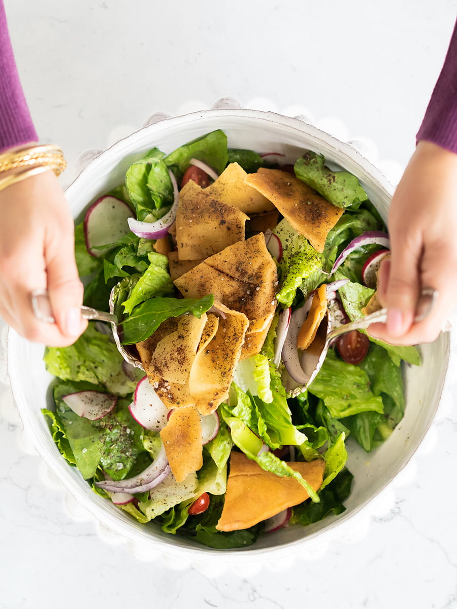 Fattoush salad in a bowl with pita chips and hands holding salad tongs to toss the salad