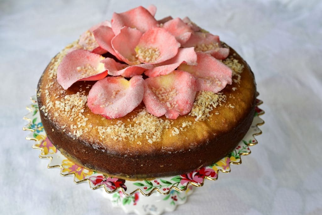 A semolina cake topped with sugared rose petals, sitting on a pedestal cake plate.