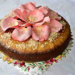 A semolina cake topped with sugared rose petals, sitting on a pedestal cake plate.